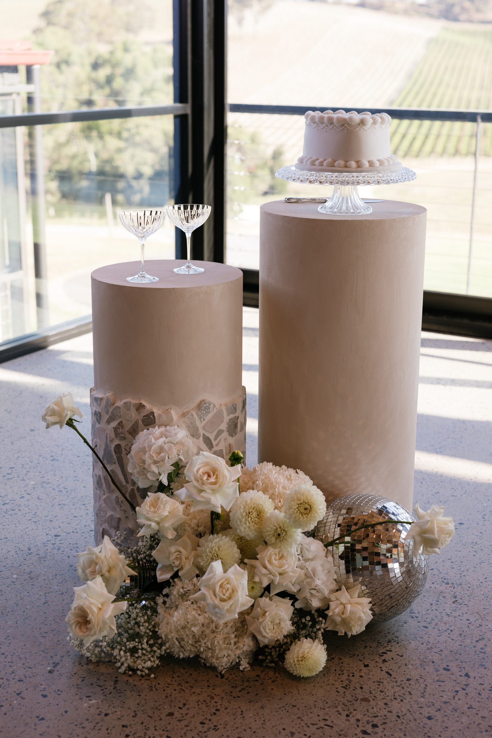 Stone plinths with white flowers, disco ball, champagne glasses and wedding cake. Styled by The Modern Approach. Photographer Jack Small