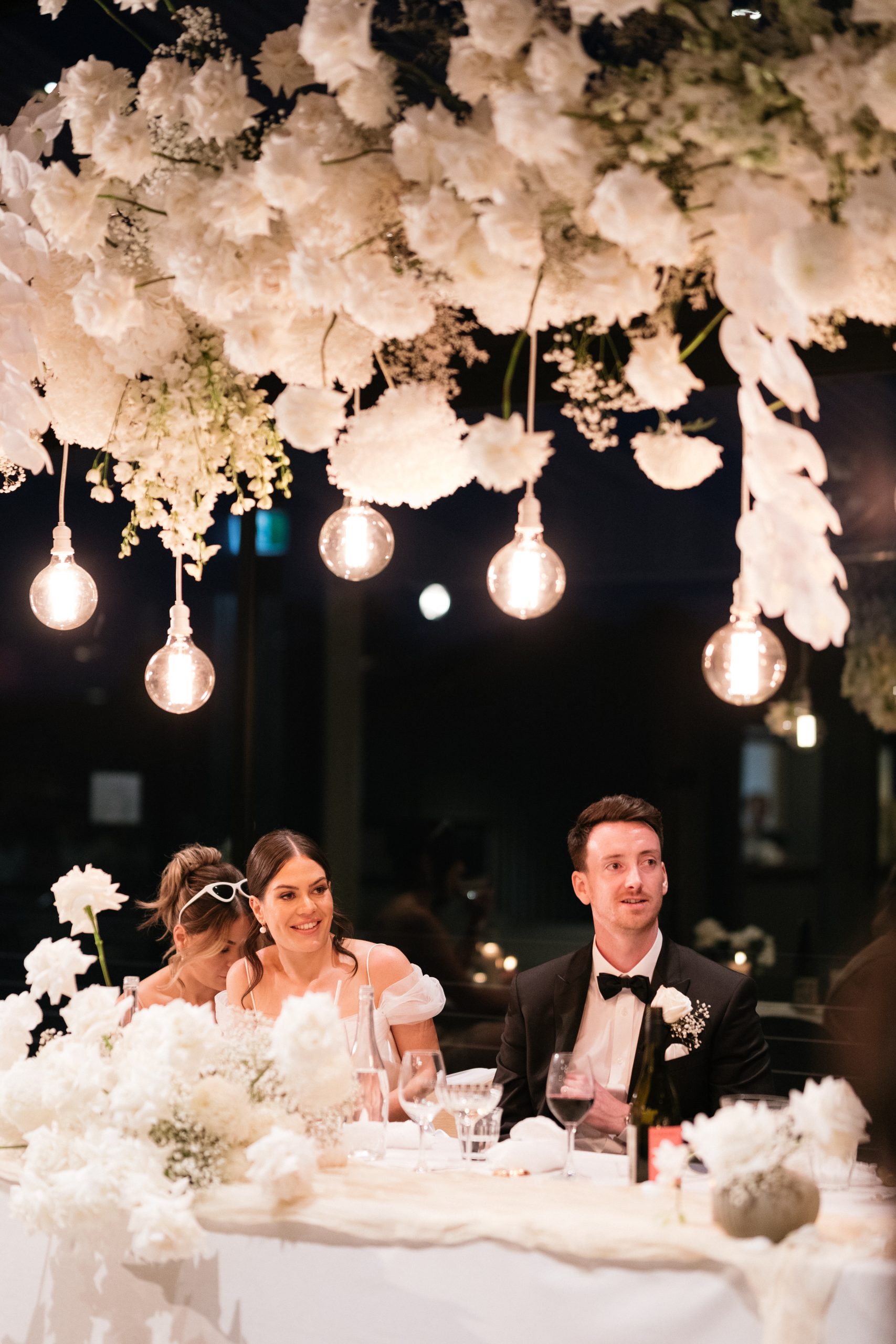 Night time reception photograph of bride and groom at wedding table with white floral overhead hanging installation and light globes