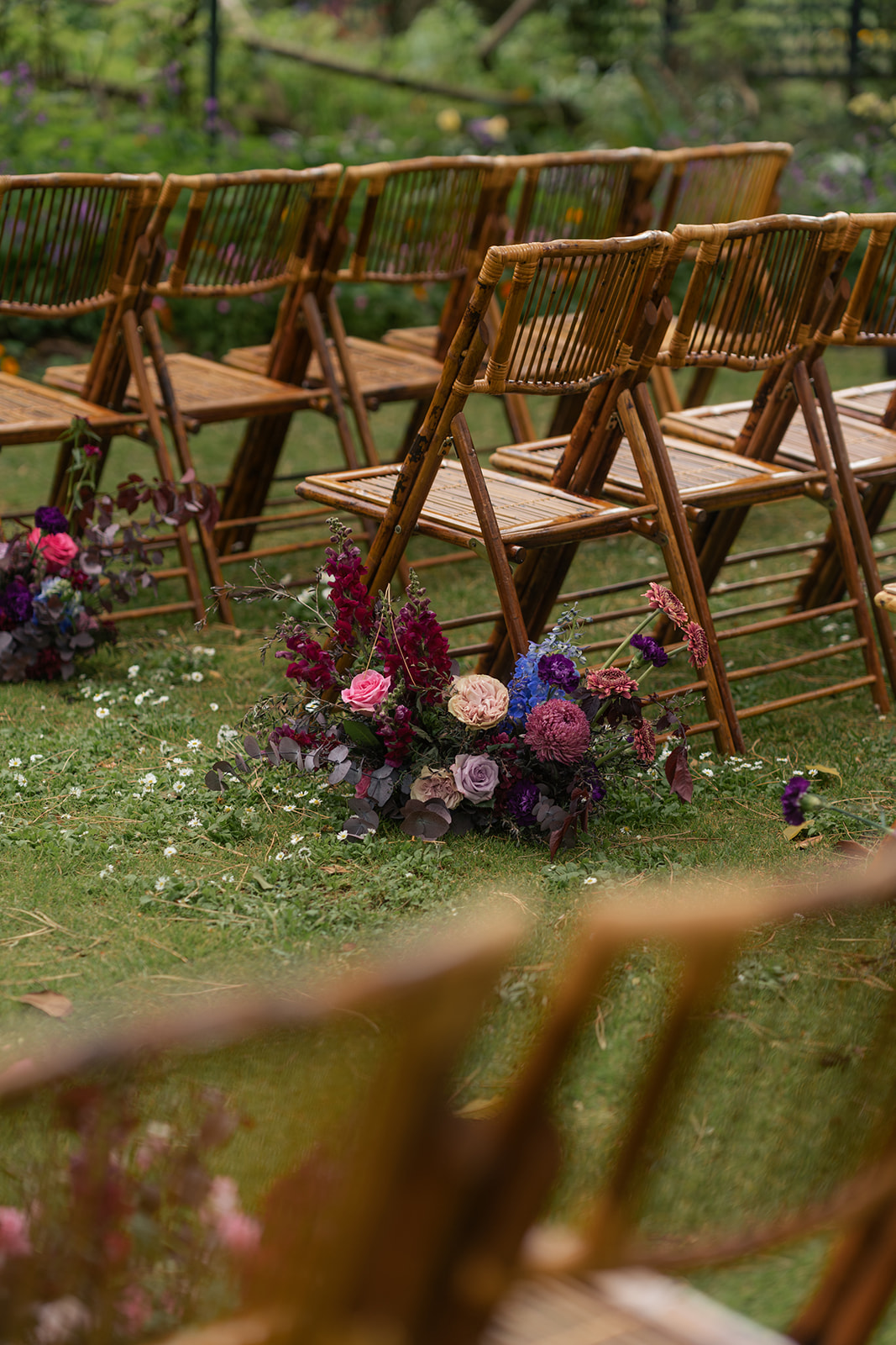 Etherial Botanica wedding florals in dark purple and pink tones lined up along chairs in the aisle to suit the couples moody rustic elegance brief
