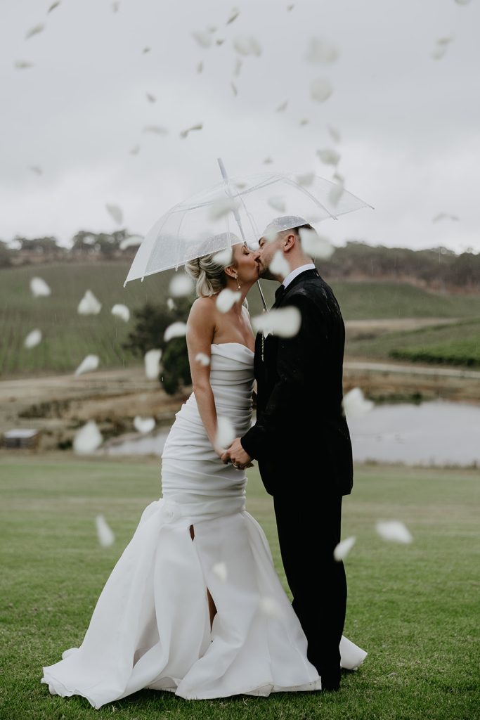 Bride and groom kissing under clear umbrella with rose petals floating in air at Longview Vineeyard