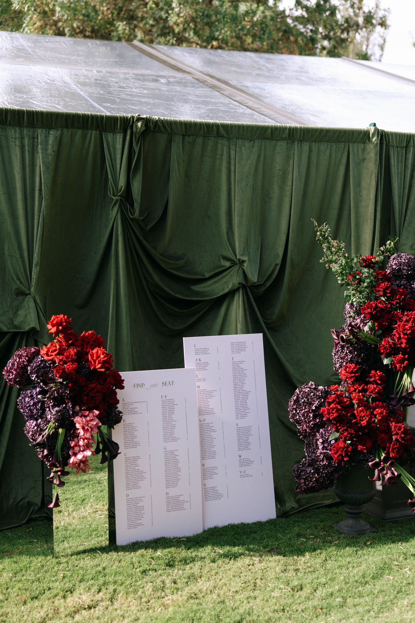 Wedding receeption seating signs displayed in front of draped green fabric with mirror and dark red and purple floral displays