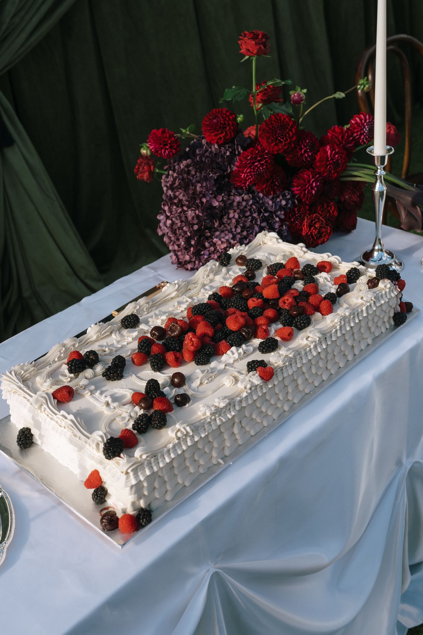 Wedding cake covered in berries displayed with dark red and purple flowers and silver candle holders