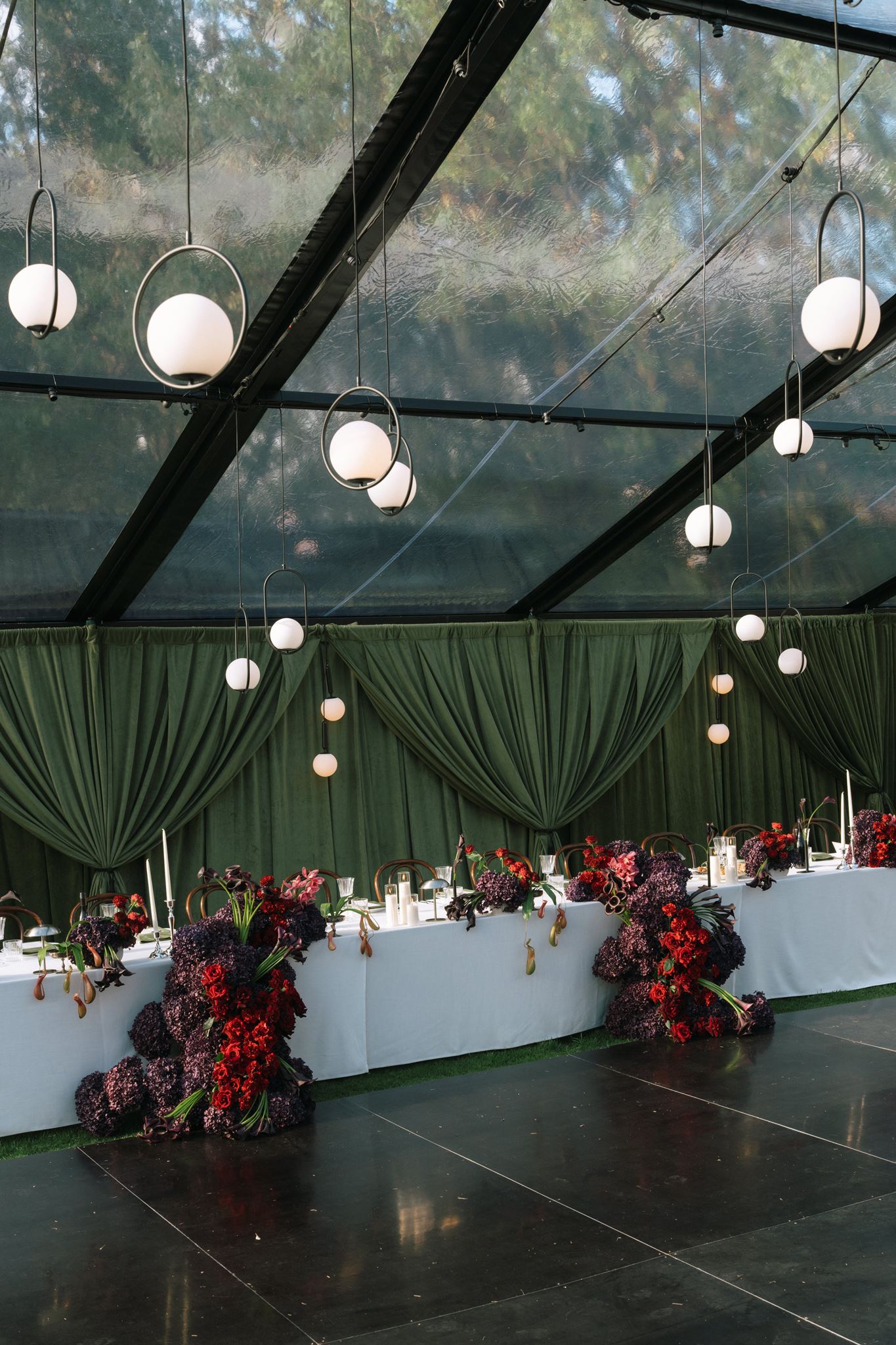 Bridal table in clear roof marquee with draped heavy greeen curtains behind and dark berry coloured floral wedding displays on table and floor