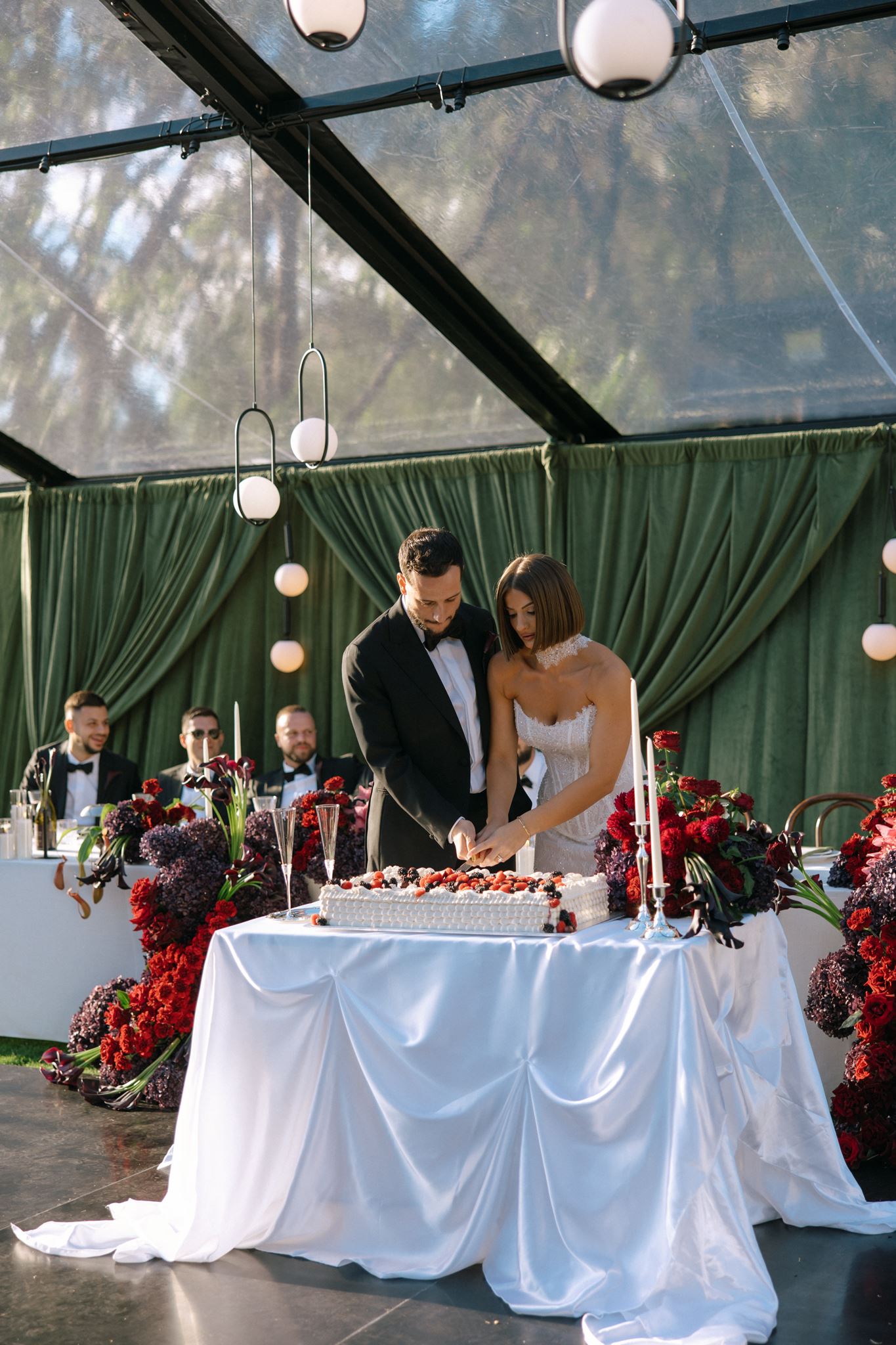 Bride and groom cutting large white and berry coloured wedding cake displayed on draped silk