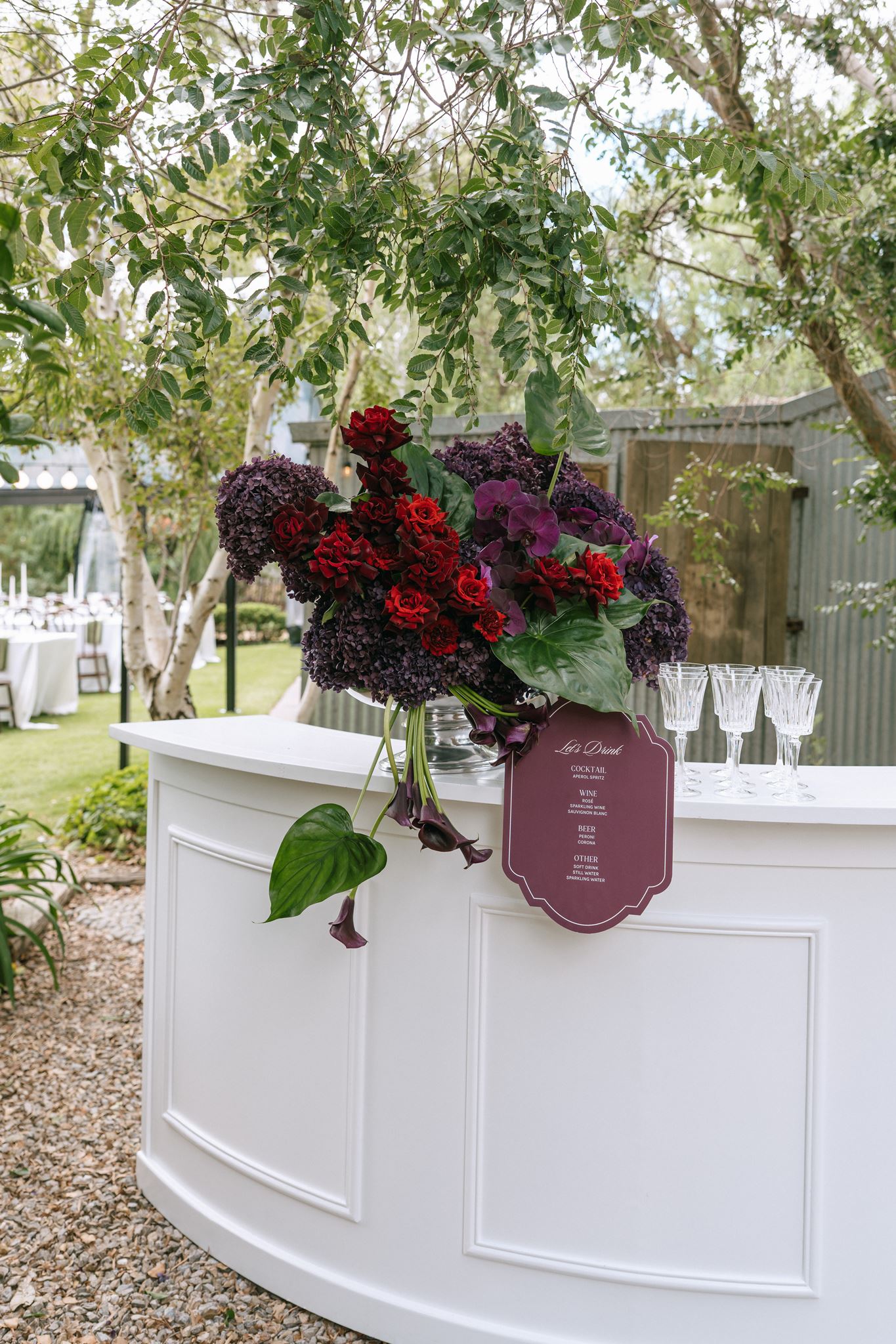 White garden bar with personalised deep purple signage and rich reds, purples and greens floral bouquet