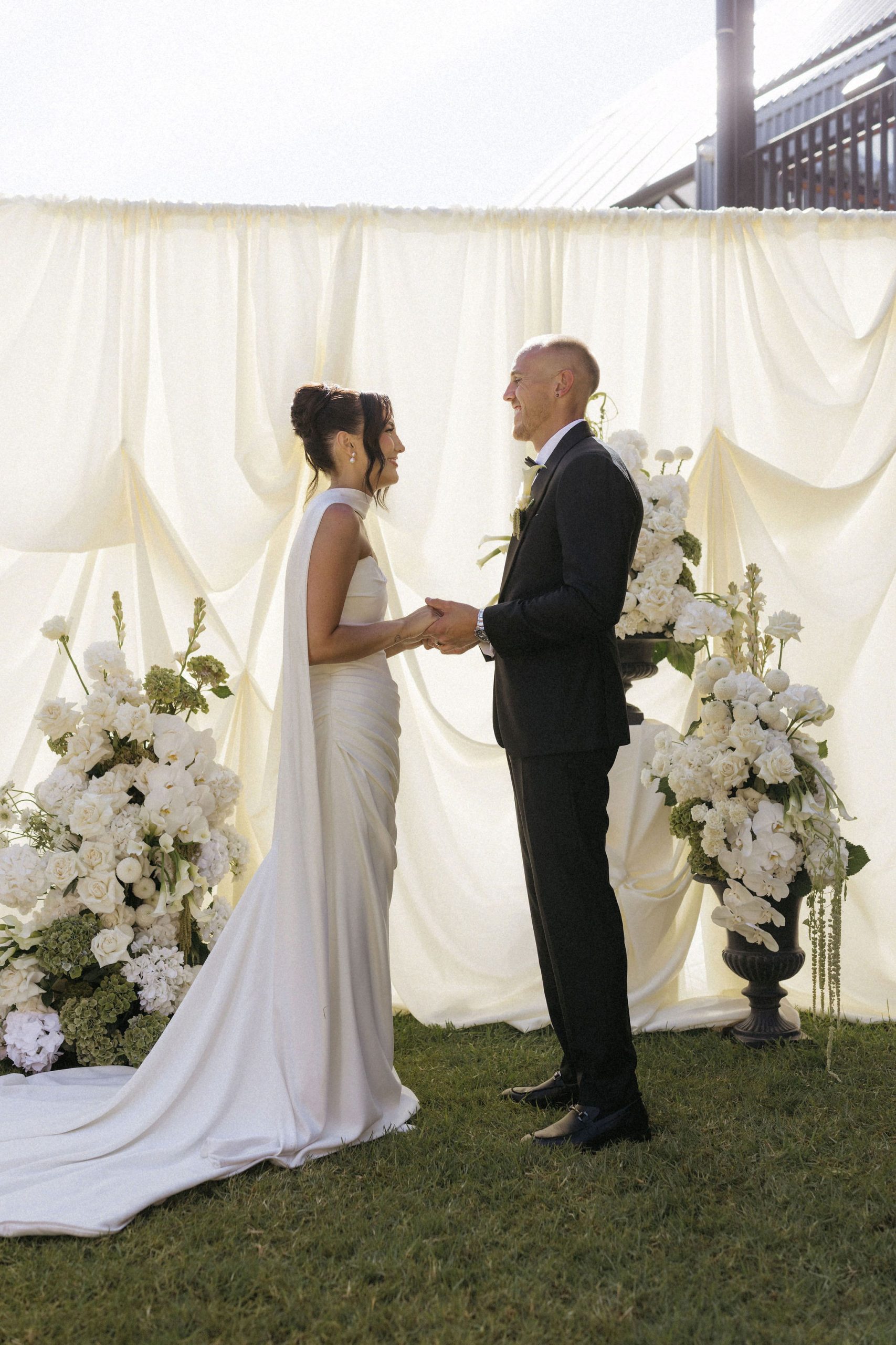 Bride and groom holding hands and standing in front of off white draped fabric arbor with large white and green modern yet classic floral floor arrangements