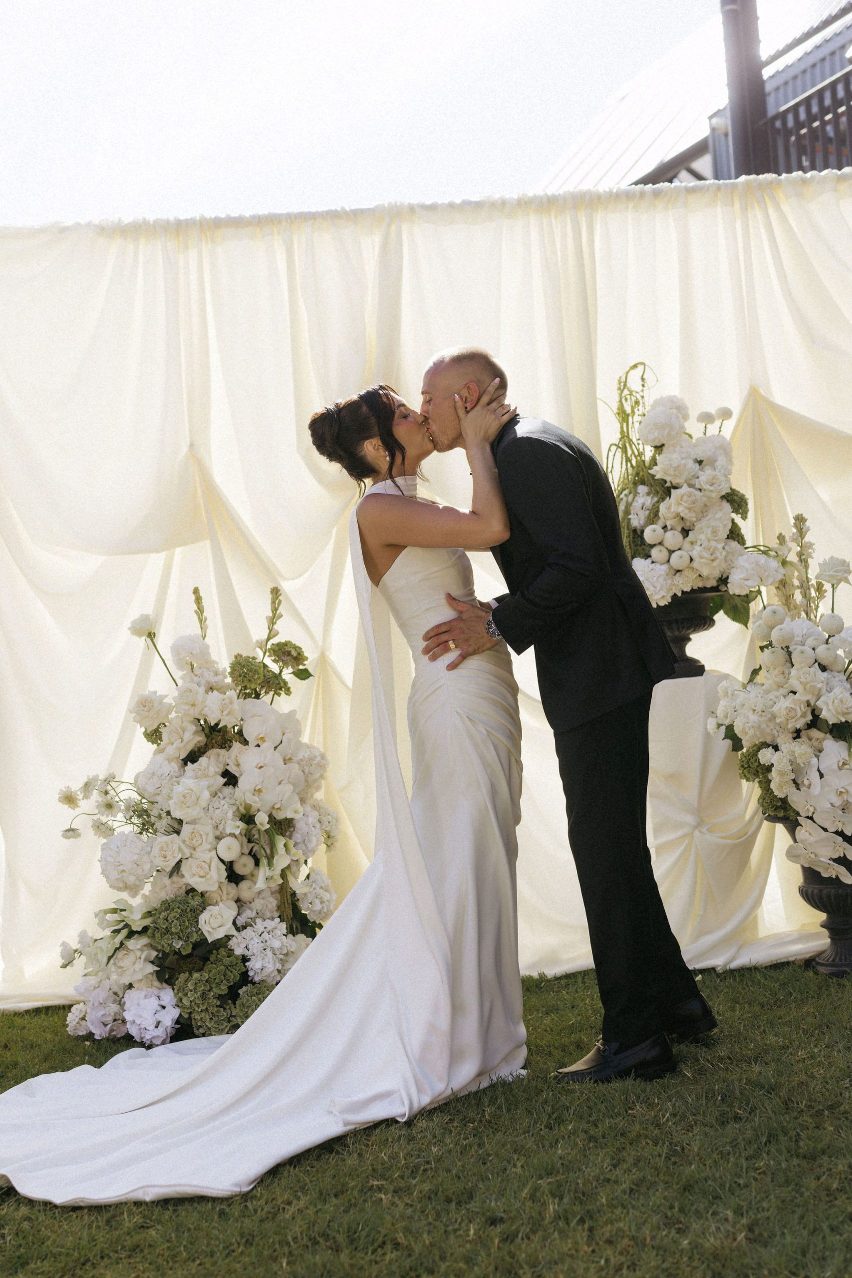 Bride and groom kissing in front of off white draped fabric arbor with large white and green modern yet classic floral floor arrangements