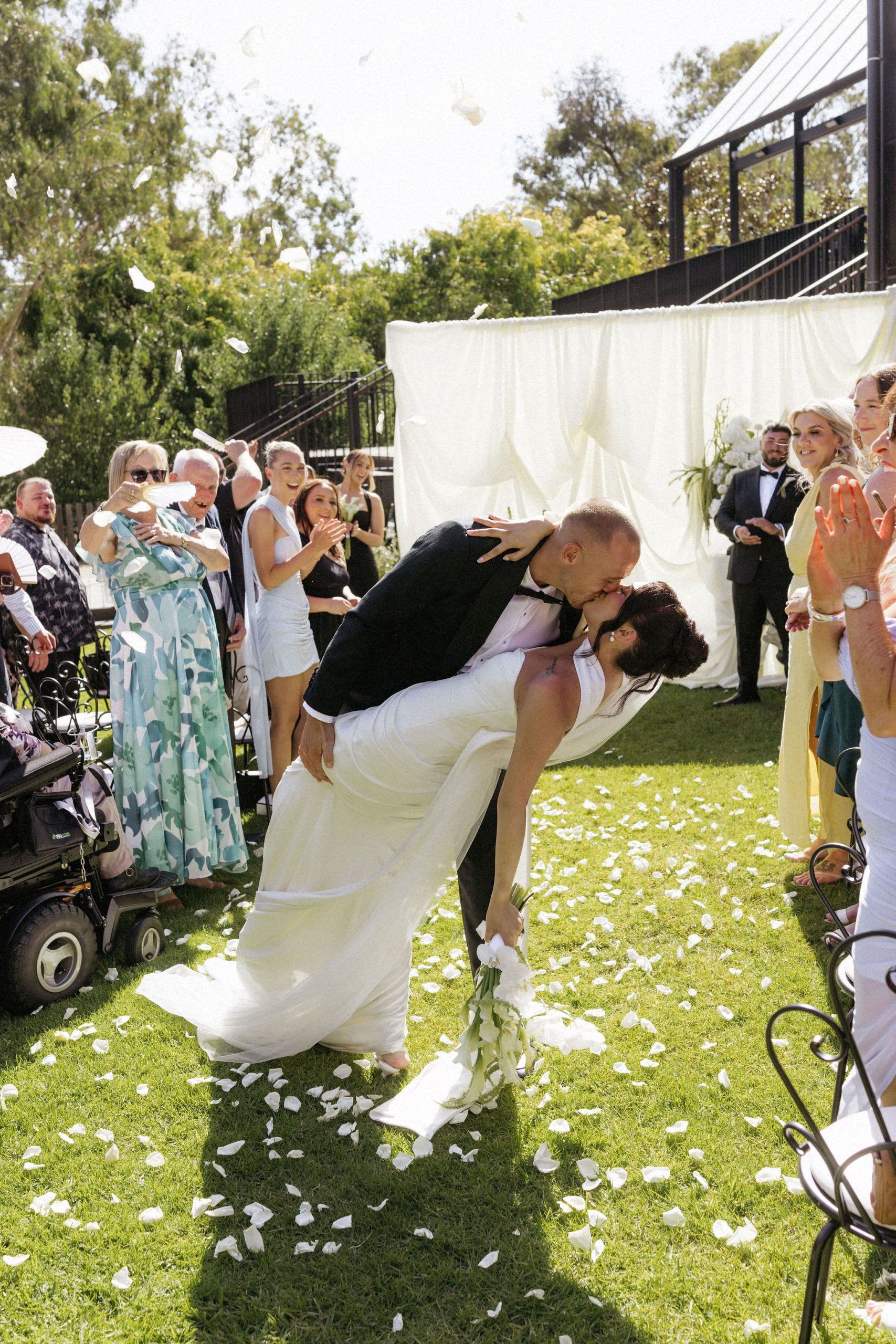 Groom bending bride backwards to kiss down the wedding aisle whilst guest throw white rose petals over them